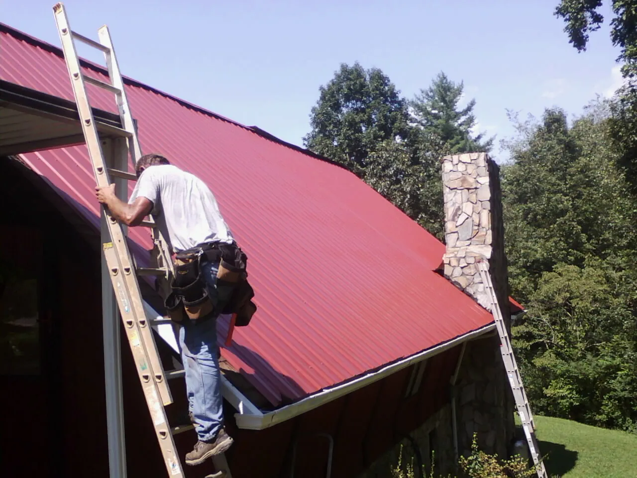Roofer on a ladder beside a red metal roof and stone chimney — Soffit & Fascia Repair by Roofing Brothers in New Bremen